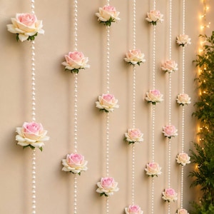 May include: A decorative hanging curtain featuring multiple vertical strands of white faux pearls interspersed with artificial pale pink and cream-coloured rose blossoms. The strands are arranged in parallel lines to create a floral backdrop.