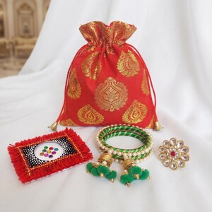 May include: A red drawstring pouch with gold patterns, a rectangular card featuring a red fringe border and a central cluster of colorful circular stickers, two green bangles with pearl accents and green pom-poms, and a floral-shaped gold ring.