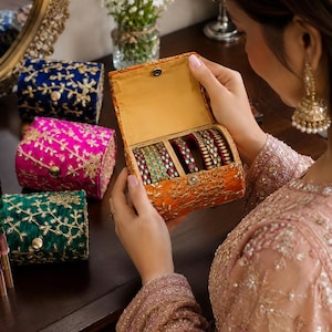 May include: A collection of rectangular fabric jewelry boxes featuring gold floral embroidery and a central snap closure. The boxes are available in orange, deep blue, bright pink, and emerald green, each containing multiple metal bangles.