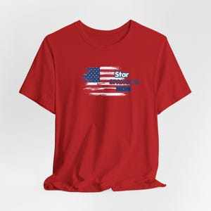 以下が含まれることがあります： 赤色のTシャツ。かすれたアメリカ国旗のデザインと、青と白で「Star Spangled Mom」の文字がプリントされています。グラフィックはシャツの前面に配置されています。