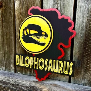 May include: A layered wooden wall sign featuring a black silhouette of a Dilophosaurus skull inside a yellow circle. Below the circle, a black rectangular plaque displays the yellow text DILOPHOSAURUS. The entire piece has a red border.