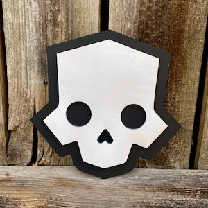 May include: A decorative skull-shaped wall hanging. The skull is white with black eye sockets and a black border. The piece is mounted on a wooden background, creating a striking contrast. This item is suitable for home decor.