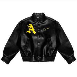 Könnte beinhalten: Schwarze Collegejacke mit gelbem "A" Emblem und goldener Schrift. Die Jacke hat einen gerippten Saum und Bündchen, einen Knopfverschluss und zwei Vordertaschen. Hergestellt aus einem lederähnlichen Material.