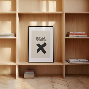 Op de afbeelding: Een ingelijste kunstdruk met het woord "BEN" boven een grote zwarte "X" op een cr&egrave;mekleurige achtergrond. Het kunstwerk wordt tentoongesteld in een licht houten boekenkast met verschillende stapels boeken.
