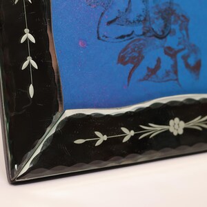 Pu&ograve; includere: Una cornice rettangolare caratterizzata da un ampio bordo nero con bordi interni smerlati e delicati motivi a tralci floreali incisi in bianco. La cornice racchiude un inserto blu brillante che mostra una tenue illustrazione botanica astratta.
