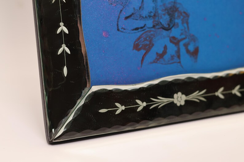 Pu&ograve; includere: Una cornice rettangolare caratterizzata da un ampio bordo nero con bordi interni smerlati e delicati motivi a tralci floreali incisi in bianco. La cornice racchiude un inserto blu brillante che mostra una tenue illustrazione botanica astratta.