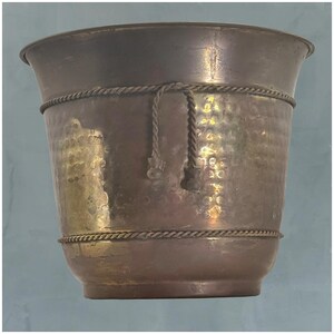 Puede incluir: Una maceta de cobre martillado con borde acampanado y detalle de cuerda. La maceta tiene una superficie texturizada y un acabado metálico cálido. Adecuada para uso en interiores o exteriores. Altura aproximada 30 cm.