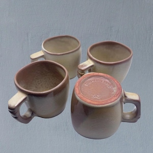 May include: Four beige ceramic mugs with brown rims and handles are arranged on a grey surface. The bottom of one mug displays the text "FRANKOMA" and "5C". The mugs have a simple, rounded design.