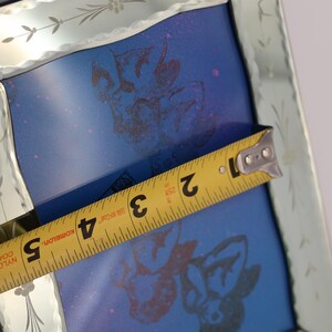 Pu&ograve; includere: Una cornice quadrata per specchio con bordo smussato e motivi floreali incisi. Un metro a nastro giallo &egrave; posizionato al centro, mostrando una larghezza di circa 12,7 cm. L'interno presenta una superficie blu con stampe grafiche scure.