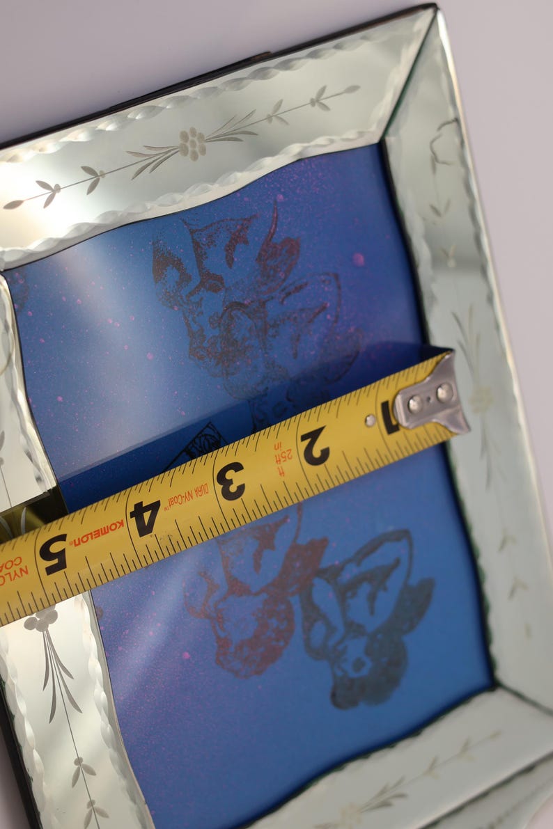 Pu&ograve; includere: Una cornice quadrata per specchio con bordo smussato e motivi floreali incisi. Un metro a nastro giallo &egrave; posizionato al centro, mostrando una larghezza di circa 12,7 cm. L'interno presenta una superficie blu con stampe grafiche scure.