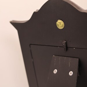 Pu&ograve; includere: Il retro di una cornice nera con un bordo superiore curvo. Un piccolo adesivo giallo circolare &egrave; visibile vicino al centro superiore con il testo MADE IN PHILIPPINES. Un supporto nero &egrave; fissato al centro con due viti argentate e una piccola chiusura.