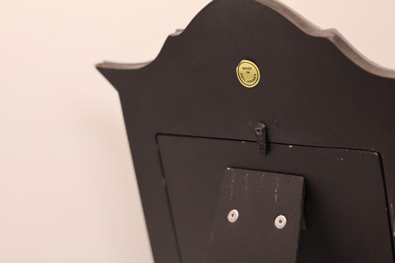 Pu&ograve; includere: Il retro di una cornice nera con un bordo superiore curvo. Un piccolo adesivo giallo circolare &egrave; visibile vicino al centro superiore con il testo MADE IN PHILIPPINES. Un supporto nero &egrave; fissato al centro con due viti argentate e una piccola chiusura.