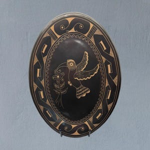 Puede incluir: Plato ovalado de cerámica negra con diseño de pájaro y flor. El pájaro sostiene una flor en el pico. El plato tiene un borde decorativo con patrones geométricos y de ondas. El pájaro y el borde son de color beige sobre fondo negro.