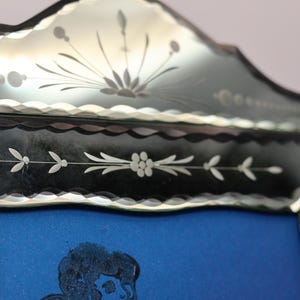 Pu&ograve; includere: Una cornice decorativa per specchio con bordo smerlato e motivi floreali incisi. Il bordo in vetro presenta complessi intagli botanici con un motivo centrale a raggiera nella parte superiore, attorno a un pannello centrale blu rettangolare.