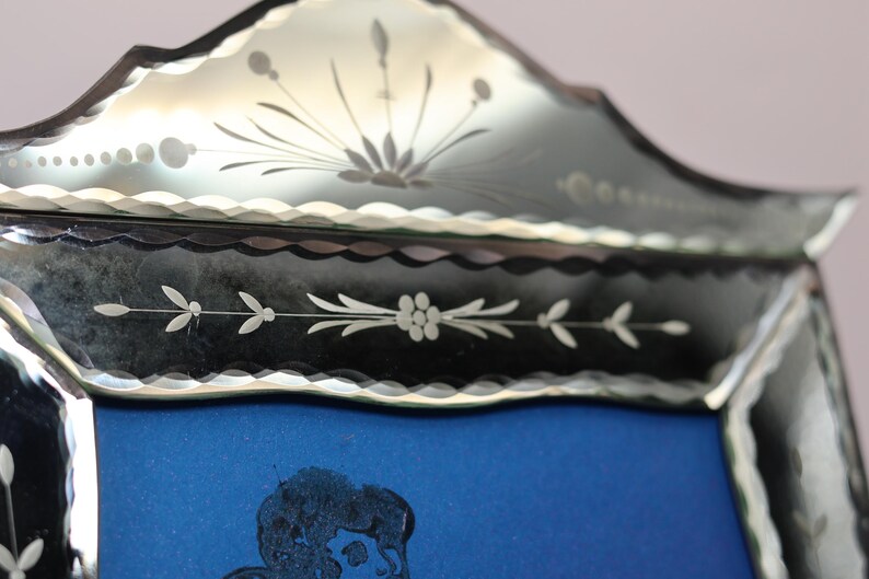 Pu&ograve; includere: Una cornice decorativa per specchio con bordo smerlato e motivi floreali incisi. Il bordo in vetro presenta complessi intagli botanici con un motivo centrale a raggiera nella parte superiore, attorno a un pannello centrale blu rettangolare.