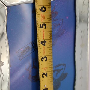 Pu&ograve; includere: Uno specchio rettangolare con una cornice in vetro bisellato e inciso con motivi floreali. Un metro a nastro giallo &egrave; posizionato verticalmente al centro, mostrando un'altezza di circa 17,8 cm. Il pannello centrale blu mostra un logo.
