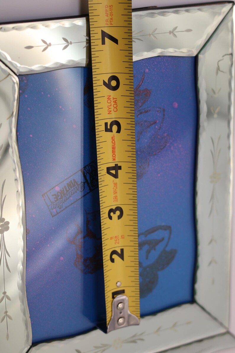 Pu&ograve; includere: Uno specchio rettangolare con una cornice in vetro bisellato e inciso con motivi floreali. Un metro a nastro giallo &egrave; posizionato verticalmente al centro, mostrando un'altezza di circa 17,8 cm. Il pannello centrale blu mostra un logo.