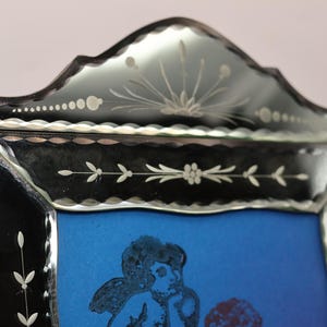 Pu&ograve; includere: Una cornice decorativa per specchio con bordo in vetro nero e motivi floreali e botanici incisi. La cornice presenta un bordo superiore smerlato e una finitura smussata, che circonda un pannello centrale blu con un'illustrazione scura.