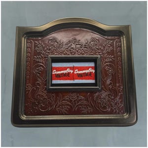 Puede incluir: Un marco rectangular marrón de estilo vintage con intrincados grabados florales y de querubines. El marco rodea un inserto rectangular más pequeño con una etiqueta roja y blanca que dice "SammyBoy VINTAGE". El marco tiene un borde de color bronce.