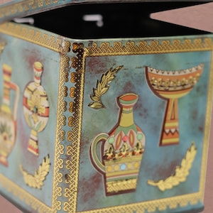 May include: A square metal tin with a hinged lid, featuring a weathered teal blue finish. The exterior is decorated with gold-toned Celtic knot borders and colorful illustrations of ancient-style pottery vessels and golden laurel leaf motifs.