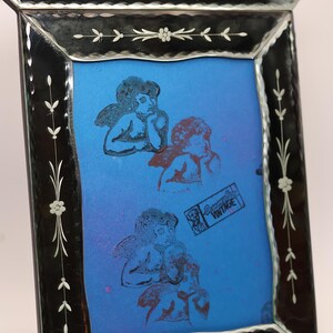 Pu&ograve; includere: Una cornice portafoto rettangolare in vetro con bordo nero caratterizzata da motivi floreali incisi e una parte superiore arcuata decorativa. La cornice contiene un inserto blu brillante che mostra tre figure di cherubini stampate e un timbro rettangolare con la scritta "Dreamy Day VINTAGE".