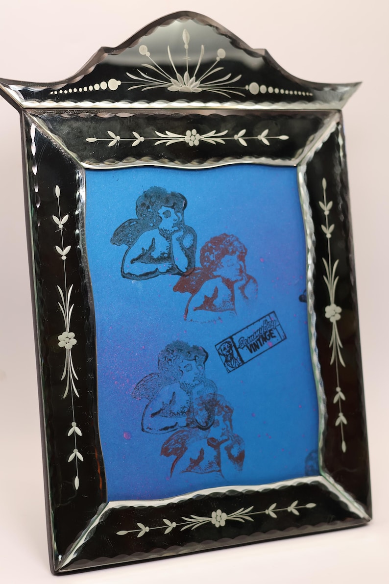 Pu&ograve; includere: Una cornice portafoto rettangolare in vetro con bordo nero caratterizzata da motivi floreali incisi e una parte superiore arcuata decorativa. La cornice contiene un inserto blu brillante che mostra tre figure di cherubini stampate e un timbro rettangolare con la scritta "Dreamy Day VINTAGE".