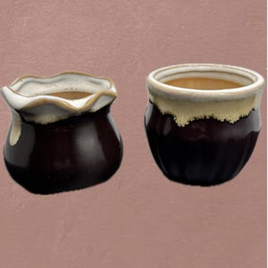 Könnte beinhalten: Zwei kleine, dunkelbraune Keramiktöpfe mit glänzendem Finish. Die Ränder sind cremeweiß mit gewelltem Rand, der Innenraum hellbraun. Ein Topf hat einen gerüschten Rand, der andere einen glatten, abgerundeten Rand.
