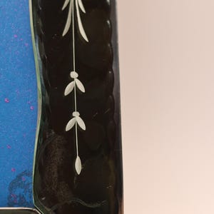 Pu&ograve; includere: Una cornice in vetro rettangolare con un bordo scuro e testurizzato dal profilo smerlato. Il bordo &egrave; decorato con un motivo floreale inciso in bianco. Un pannello blu vibrante con sottili macchie viola &egrave; visibile sul lato sinistro della cornice.