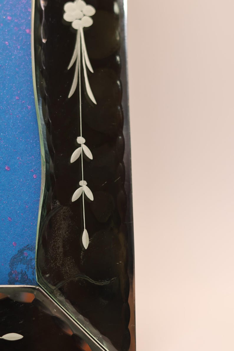 Pu&ograve; includere: Una cornice in vetro rettangolare con un bordo scuro e testurizzato dal profilo smerlato. Il bordo &egrave; decorato con un motivo floreale inciso in bianco. Un pannello blu vibrante con sottili macchie viola &egrave; visibile sul lato sinistro della cornice.