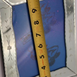 Pu&ograve; includere: Una cornice per specchio rettangolare con bordi smussati e motivi floreali incisi. Un metro a nastro giallo &egrave; posizionato verticalmente al centro, mostrando una lunghezza di circa 26,7 centimetri dal bordo superiore a quello inferiore.