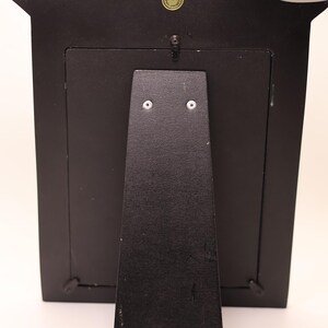 Pu&ograve; includere: Una cornice rettangolare nera vista dal retro, dotata di un supporto centrale fissato con due viti argentate, una piccola clip girevole in metallo nella parte superiore e un adesivo circolare vicino al bordo superiore con la scritta Made in Philippines.