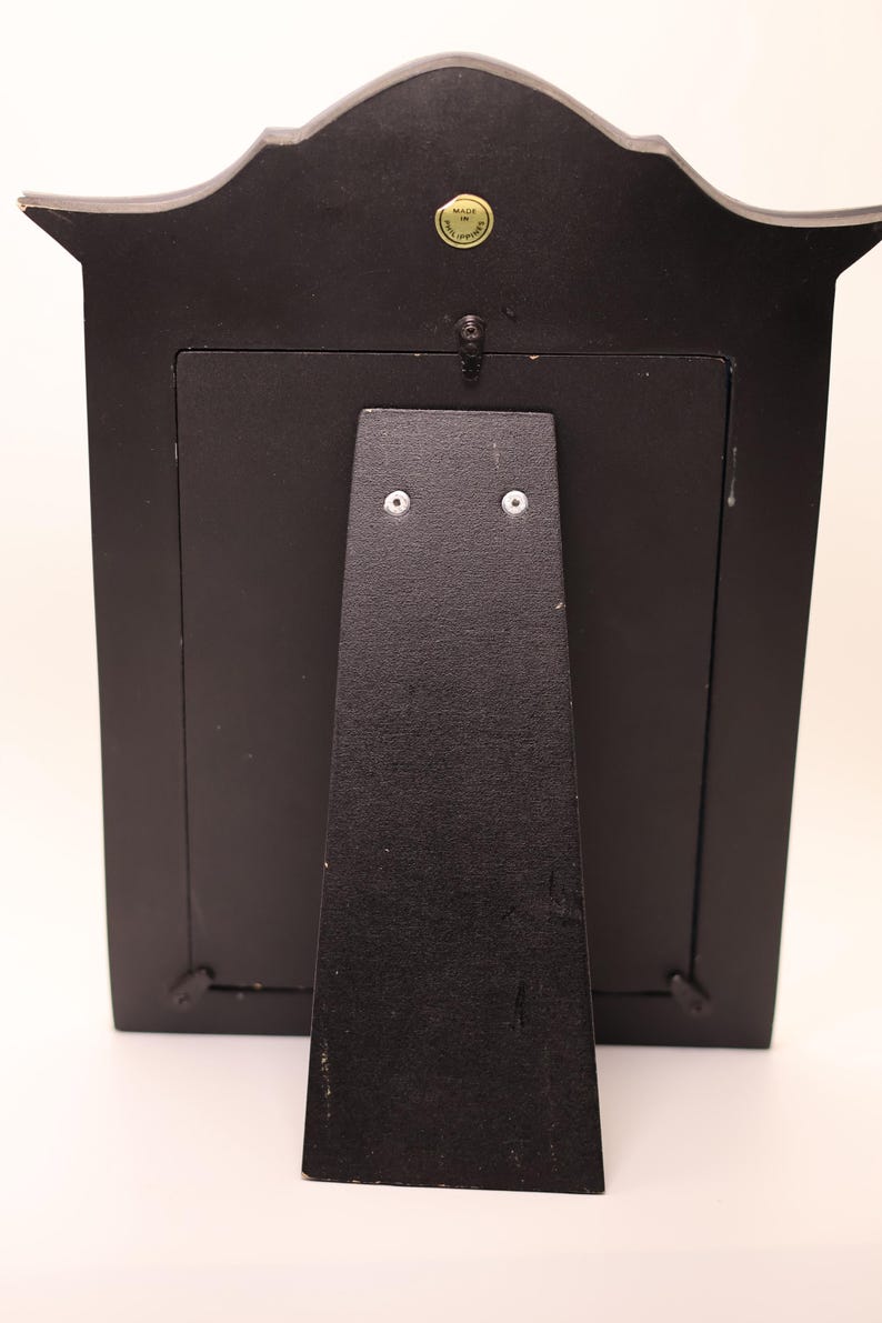 Pu&ograve; includere: Una cornice rettangolare nera vista dal retro, dotata di un supporto centrale fissato con due viti argentate, una piccola clip girevole in metallo nella parte superiore e un adesivo circolare vicino al bordo superiore con la scritta Made in Philippines.