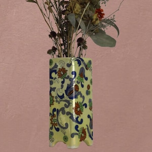 May include: A rectangular yellow vase with a floral design in blue, orange, and green. The vase has a square base with three small feet and is filled with dried flowers and branches. The background is a solid pink color.