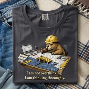 May include: Dark gray t-shirt featuring a cartoon beaver wearing a yellow hard hat, working on blueprints with a laptop and calculator. The text "I am not overthinking. I am thinking thoroughly." is printed below the beaver.