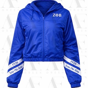 May include: A royal blue hooded jacket with a full zipper closure. The jacket features white text on the chest that reads "ZΦB". The sleeves have white stripes with the text "1920 Zeta Phi Beta".
