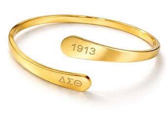 Pode incluir: Uma pulseira de punho aberta dourada. A pulseira tem o texto gravado "1913" em uma extremidade e "ΔΣΘ" na outra. A joia está em um fundo branco.