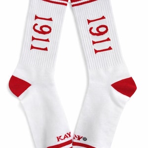 Pode incluir: Par de meias brancas com detalhes vermelhos. As meias têm dedos e calcanhares vermelhos e uma faixa vermelha na parte superior com duas listras brancas. O número "1911" é impresso em vermelho na lateral de cada meia. A marca "KAY" é impressa perto do tornozelo.