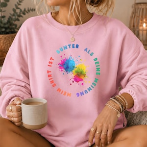 Könnte beinhalten: Ein hellrosa Sweatshirt mit Rundhalsausschnitt und einem kreisförmigen Aufdruck mit dem Text "MEIN SHIRT IST BUNTER ALS DEINE MEINUNG", der ein lebendiges, mehrfarbiges Farbspritzer-Design in Blau, Pink, Gelb und Grün umgibt.