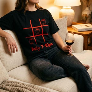 Op de afbeelding: Een zwart T-shirt met korte mouwen voorzien van een rode boter-kaas-en-eieren rasterafbeelding met wijnglaspictogrammen in de vakjes en de rode tekst Daily 7-10pm onder het raster. Het shirt wordt gecombineerd met een donkergrijze spijkerbroek.