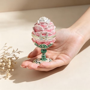Op de afbeelding: Een decoratieve sieradendoos in de vorm van een bloemknop, vastgehouden in een hand. De doos is roze en wit met gouden accenten en een juwelen strik. De basis is groen en goud, en de doos is ongeveer 7.6 cm hoog.