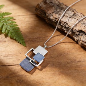 May include: Silver-toned necklace with a geometric pendant featuring squares in silver, light blue, and dark blue. The pendant is connected to a delicate chain. The necklace is displayed on a wooden surface.