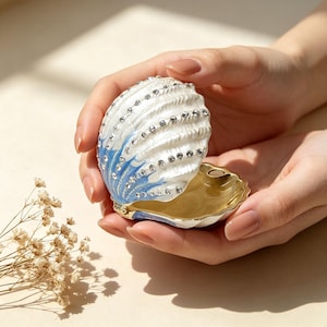 May include: A decorative seashell-shaped trinket box. The hinged box is white with blue accents and embellished with rhinestones. The interior is gold-colored. The box is held open in a person's hands.