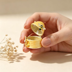 May include: A yellow and gold decorative trinket box shaped like a beehive, with a hinged lid. The box features a small, detailed bee on the lid and a honey-colored interior. The box is held open, showcasing its contents.