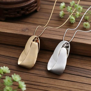 Puede incluir: Dos collares con diseños de colgantes. Uno presenta un colgante dorado, mientras que el otro tiene un colgante plateado. Ambos colgantes tienen una forma curva única. Los collares descansan sobre una superficie de madera.