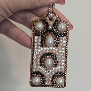 Pode incluir: Um porta-chaves retangular adornado com uma borda dourada. O design apresenta pérolas brancas, cristais pretos e transparentes e uma pérola grande no centro. Uma corrente prateada está presa no topo.