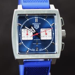 May include: A square-faced Monaco TAG Heuer watch with a blue dial and a blue rubber strap. The watch features white subdials, red hands, and the word "AUTOMATIC". The watch case is silver.