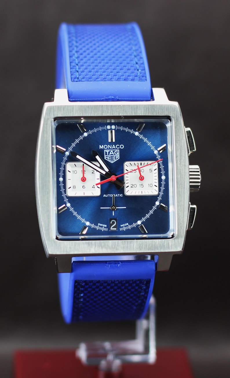 May include: A square-faced Monaco TAG Heuer watch with a blue dial and a blue rubber strap. The watch features white subdials, red hands, and the word "AUTOMATIC". The watch case is silver.