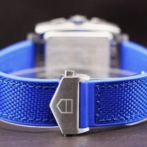 May include: Close-up of a blue watch strap with a woven texture. The strap is secured with a silver clasp. The watch face is visible in the background. The watch band is made of a blue rubber material.
