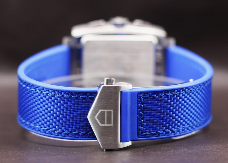May include: Close-up of a blue watch strap with a woven texture. The strap is secured with a silver clasp. The watch face is visible in the background. The watch band is made of a blue rubber material.