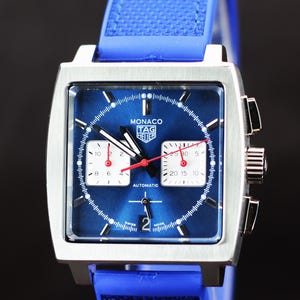 May include: A square-faced wristwatch with a blue dial and a blue rubber strap. The watch features white subdials, red hands, and the text "MONACO TAG HEUER AUTOMATIC". The watch is made in Switzerland.